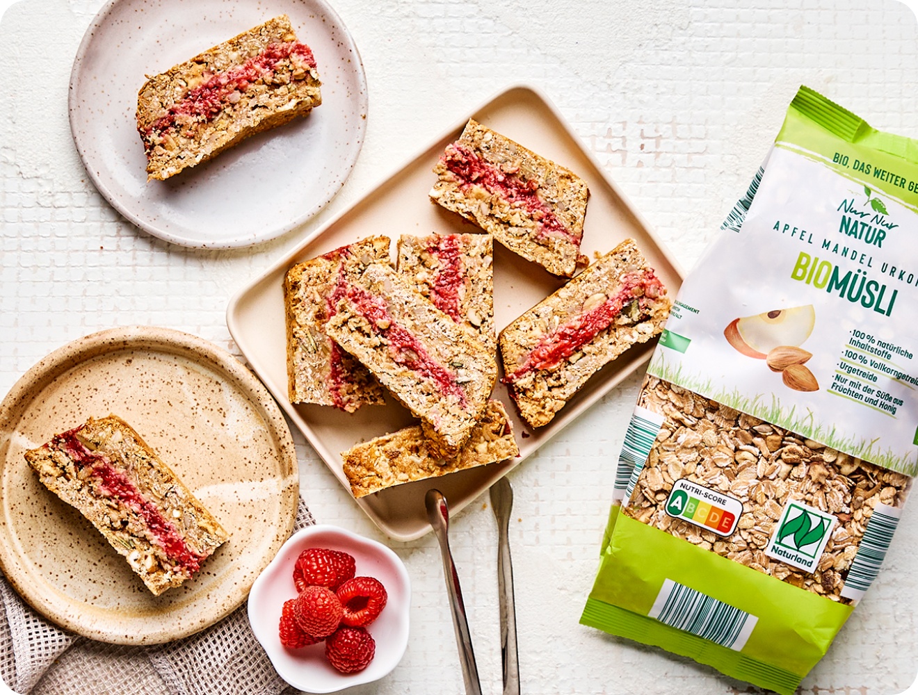 Bio-Müsliriegel mit Himbeerfüllung auf Tellern, daneben eine Packung Apfel-Mandel-Urkorn-Biomüsli und frische Himbeeren.
