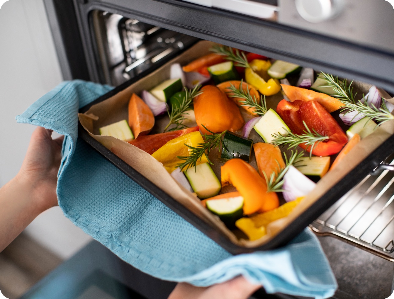 Buntes Gemüse auf einem Backblech wird in den Ofen geschoben. Auf dem Blech Rosmarin, Paprika, Zucchini und Zwiebeln.