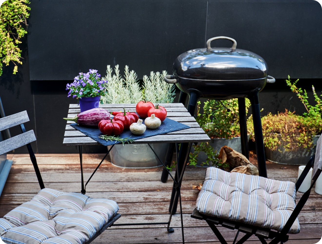 Gemüseauswahl mit Tomaten, Aubergine und Pilzen auf Holztisch vor Kugelgrill auf bepflanzter Terrasse.