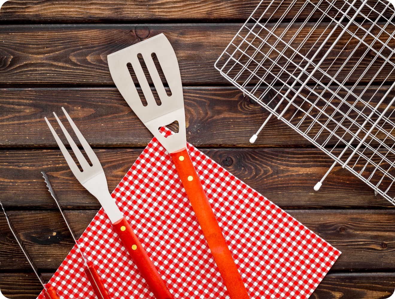 Grillbesteck mit Gabel, Zange und Wender auf rot-weißem Tuch, daneben Grillrost auf Holzuntergrund.