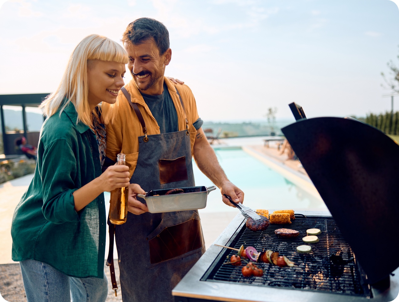 Lächelndes Paar beim Grillen am Pool, Mann in Schürze wendet Burger, Gemüse und Mais auf dem Grill.
