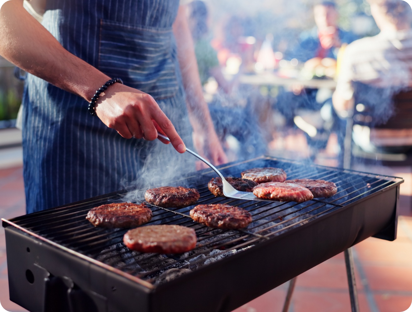 Person in gestreifter Schürze grillt mehrere Burger auf Holzkohlegrill, Rauch steigt auf, Gäste im Hintergrund.