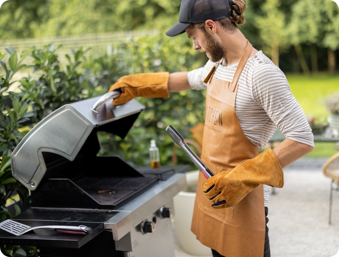 Mann mit Lederhandschuhen und Schürze öffnet Gasgrilldeckel im Garten, Grillzange in der Hand, bereit zum Grillen.