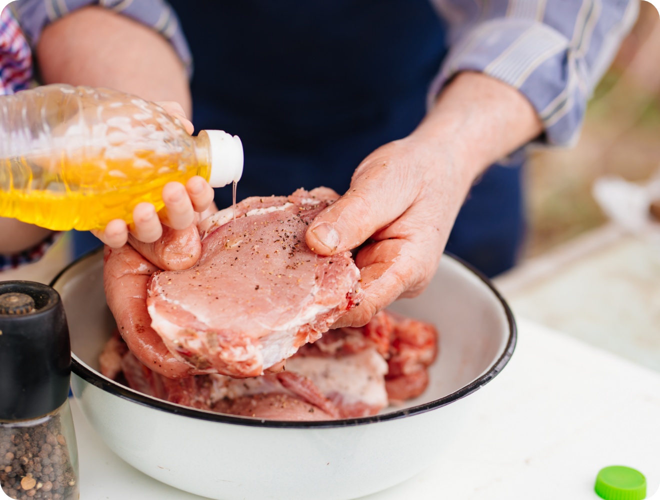Rohes Schweinefleisch wird mit Öl beträufelt und gewürzt, Hände halten Fleisch über Schüssel, Pfeffermühle daneben.