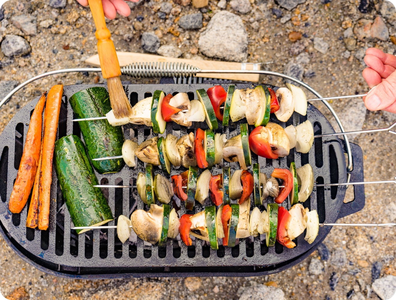 Gemüsespieße mit Paprika, Zucchini, Zwiebeln und Pilzen auf Grillrost, Möhren daneben, Marinade wird aufgetragen.