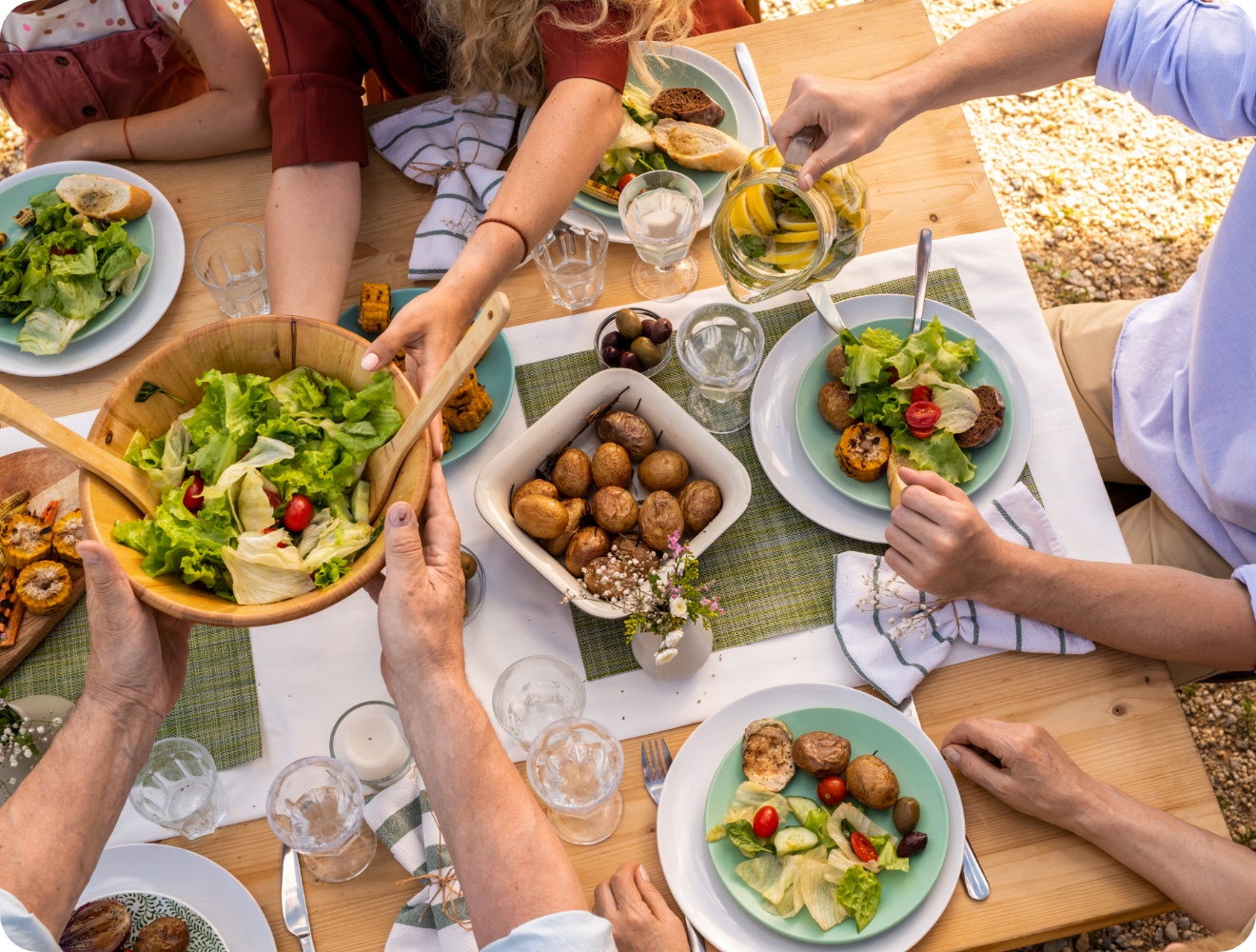Sommerliche Tischszene mit Grillgemüse, Salat, Kartoffeln und Brot, mehrere Hände greifen zu, Wasser mit Zitrone wird eingeschenkt.