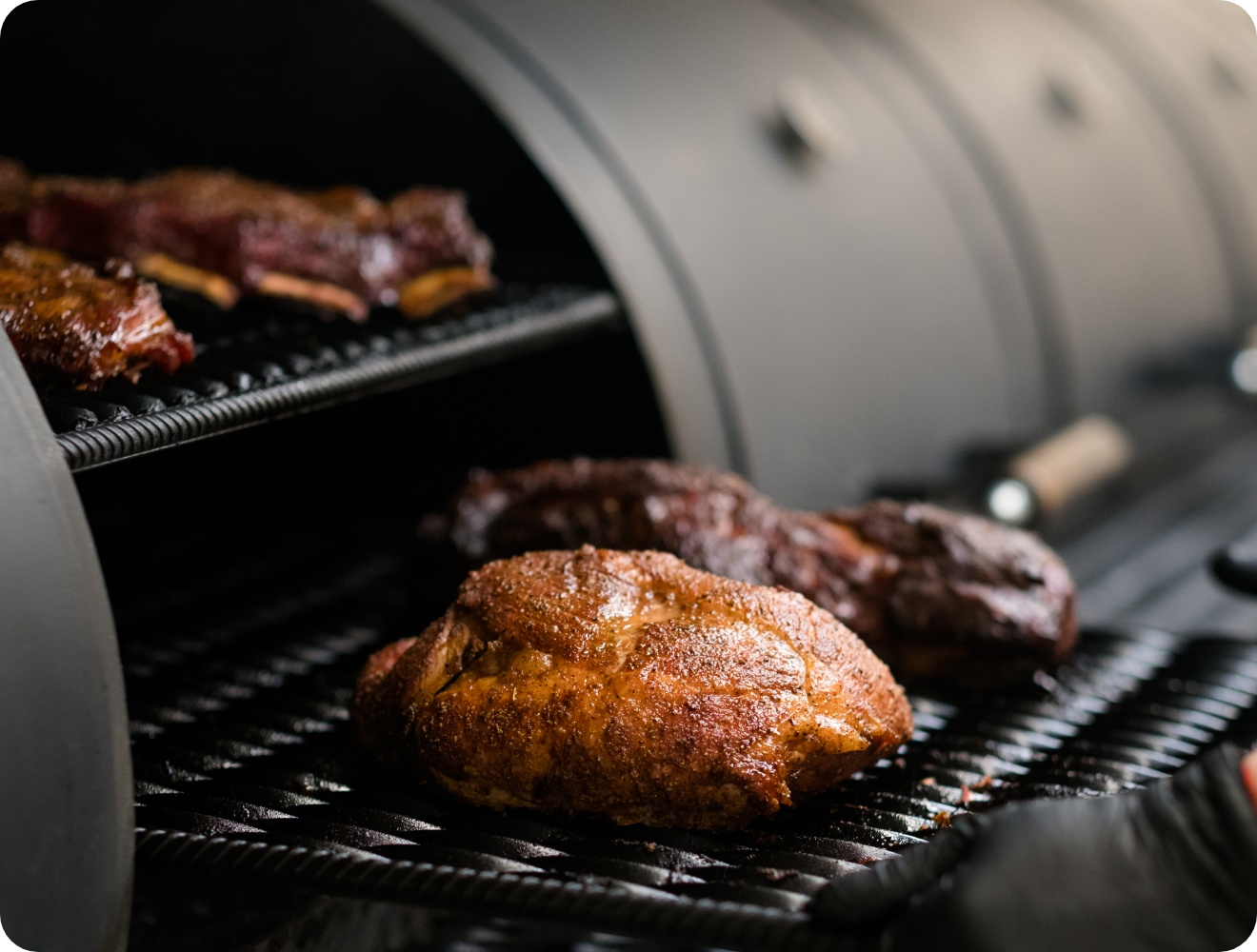 Großes Stück Fleisch mit Gewürzkruste im Smoker gegrillt, saftig gebräunt, weitere Fleischstücke auf oberem Rost.