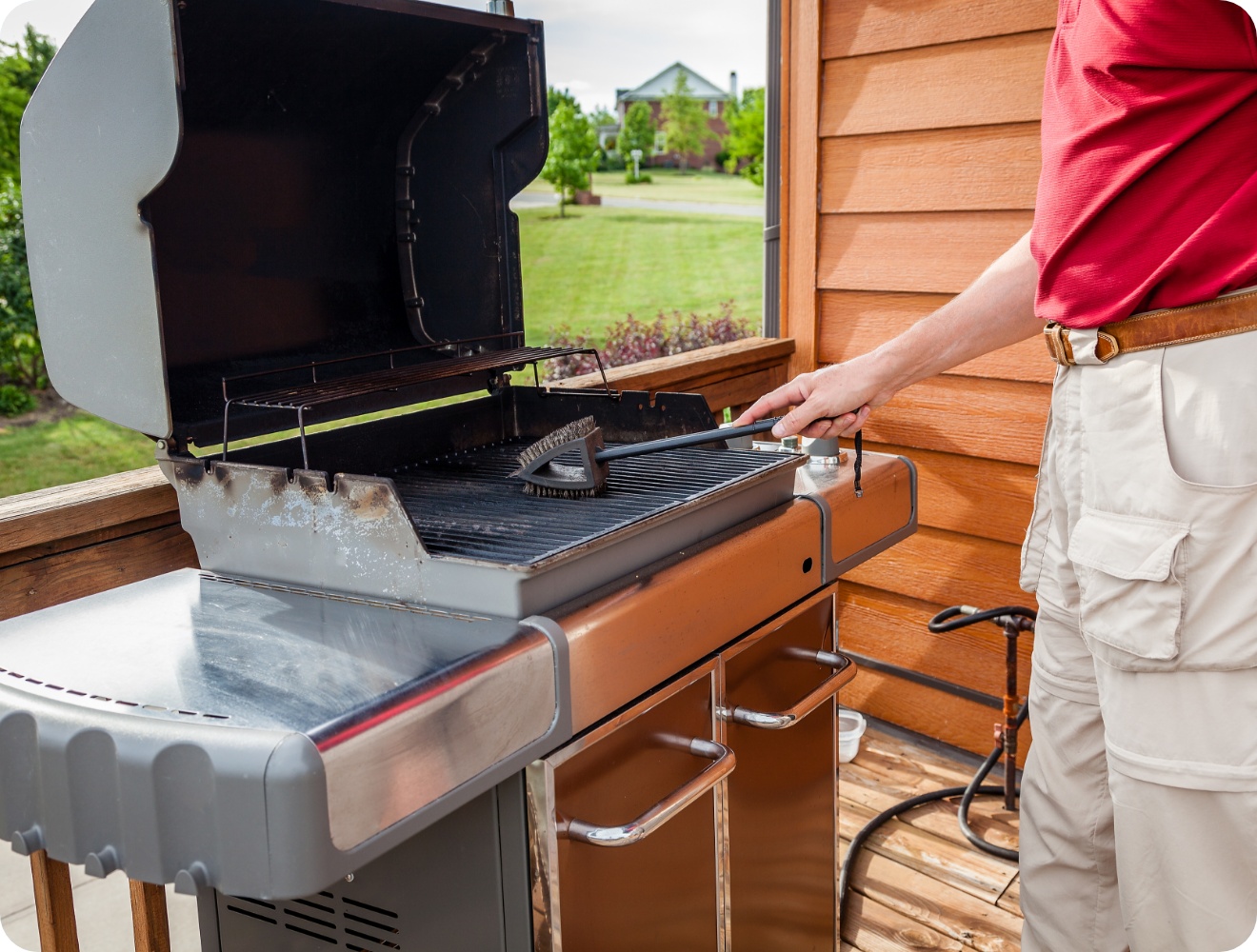 Mann reinigt Gasgrill mit Grillbürste auf Holzterrasse, Grilldeckel offen, gepflegter Garten im Hintergrund.