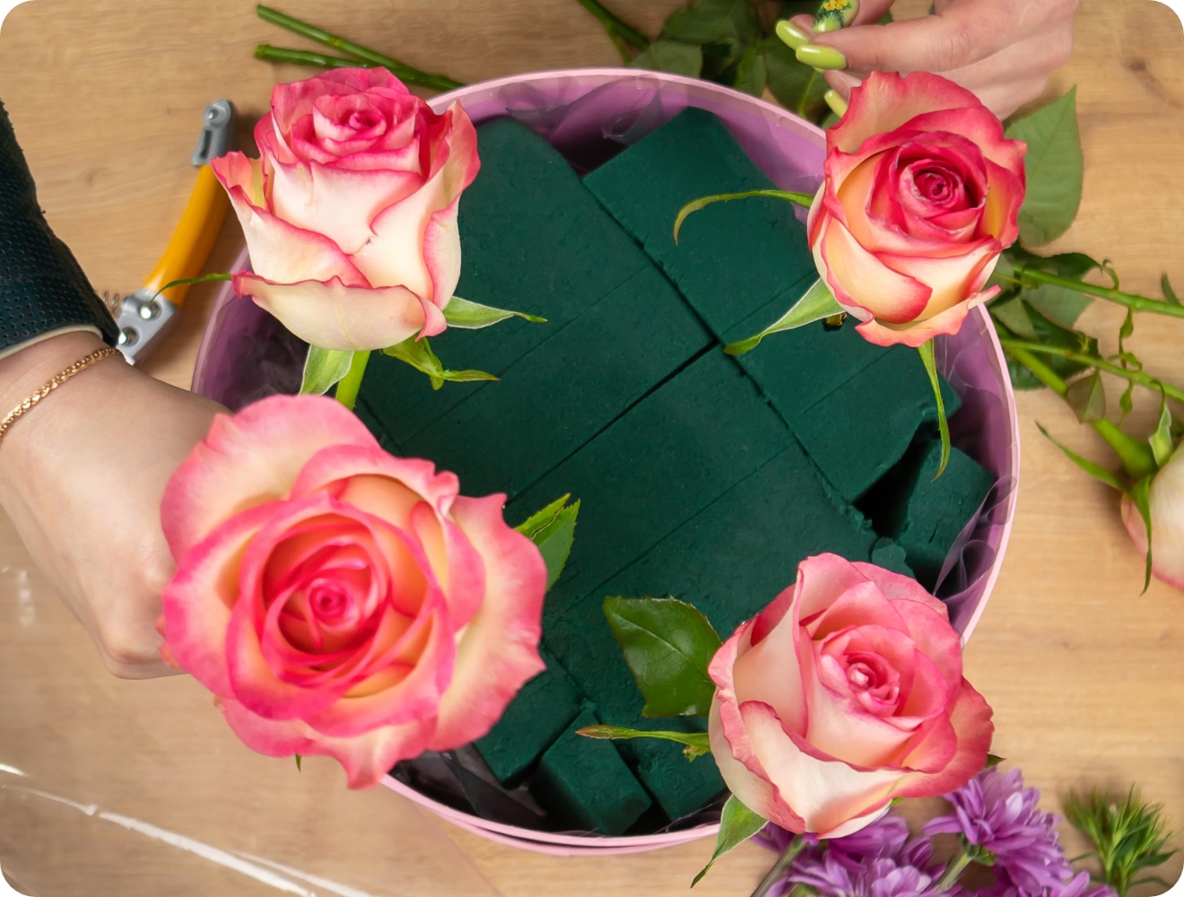 Person steckt vier rosa Rosen in einen Kreis mit grünem Steckschaum.