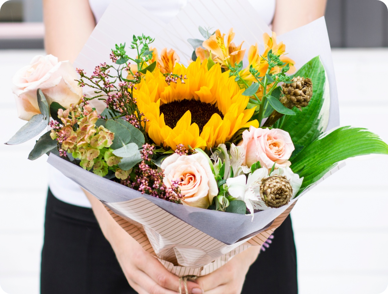 Person hält einen Blumenstrauß mit Rosen, Sonnenblumen und Grünzeug in der Hand.