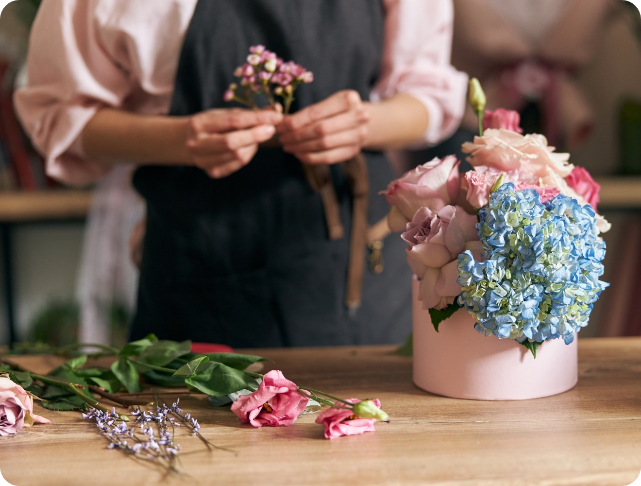 Floristin arrangiert zarte rosa Rosen und blaue Hortensien in pastellfarbener Hutschachtel auf Holztisch..