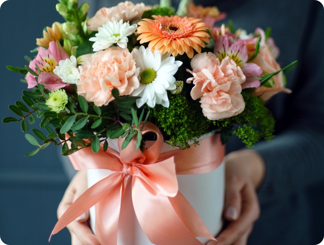 Runder Blumenstrauß mit pastellfarbenen Nelken, weißen Margeriten und rosa Schleife in Hutschachtel.