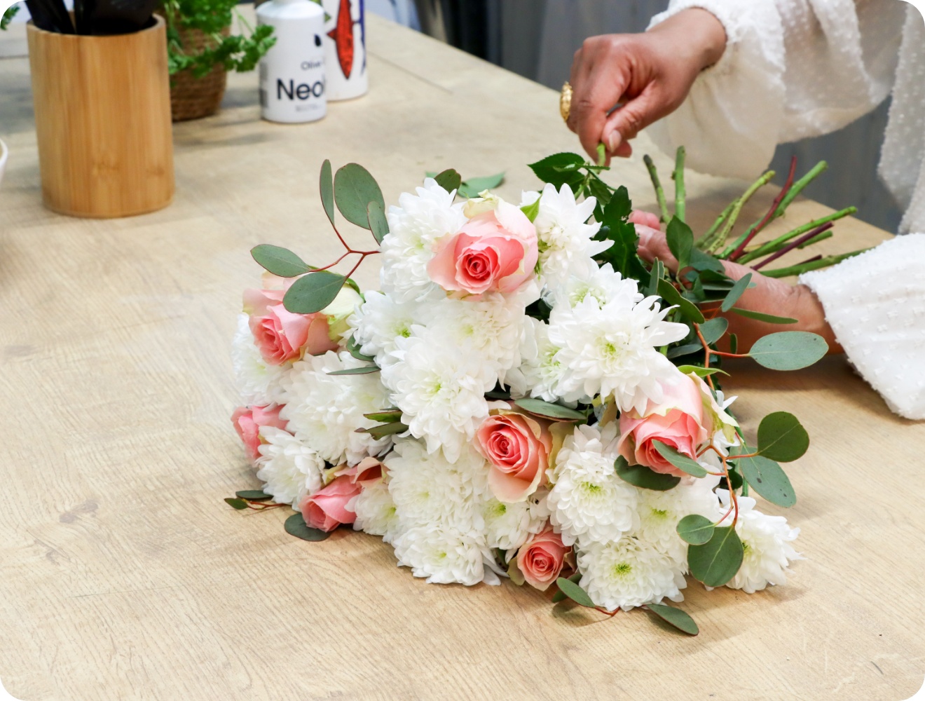 Ein gebundener Blumenstrauß aus weißen Chrysanthemen, rosa Rosen und Eucalytptus liegt auf einer Tischplatte.