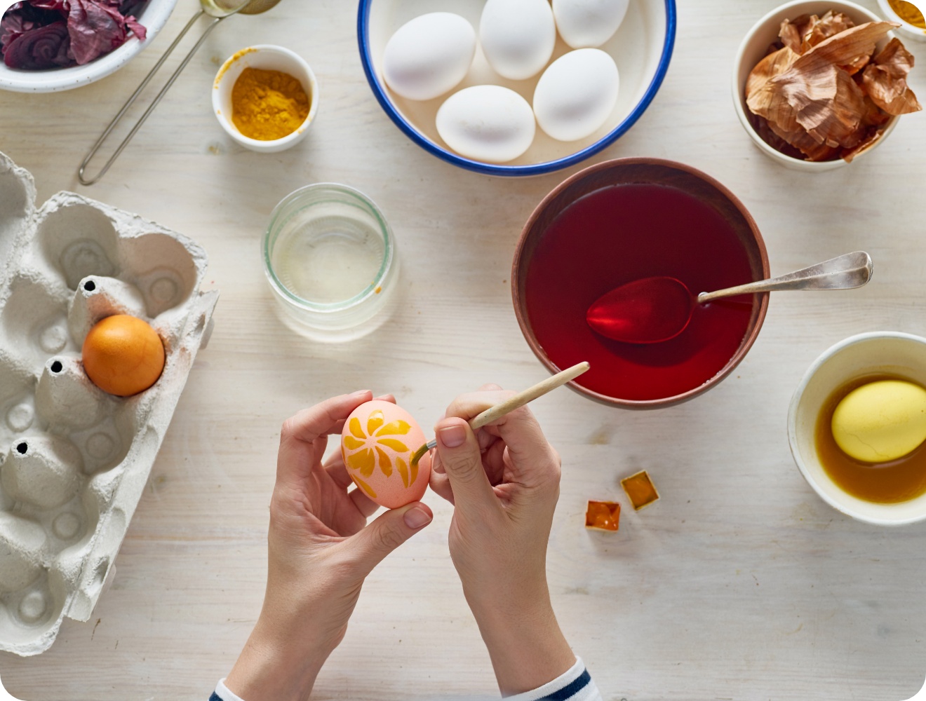 Person verziert ein Osterei mit einem floralen Muster. Auf einer weißen Arbeitsplatte stehen Kurkuma und Zwiebelschalen zum natürlichen Färben.