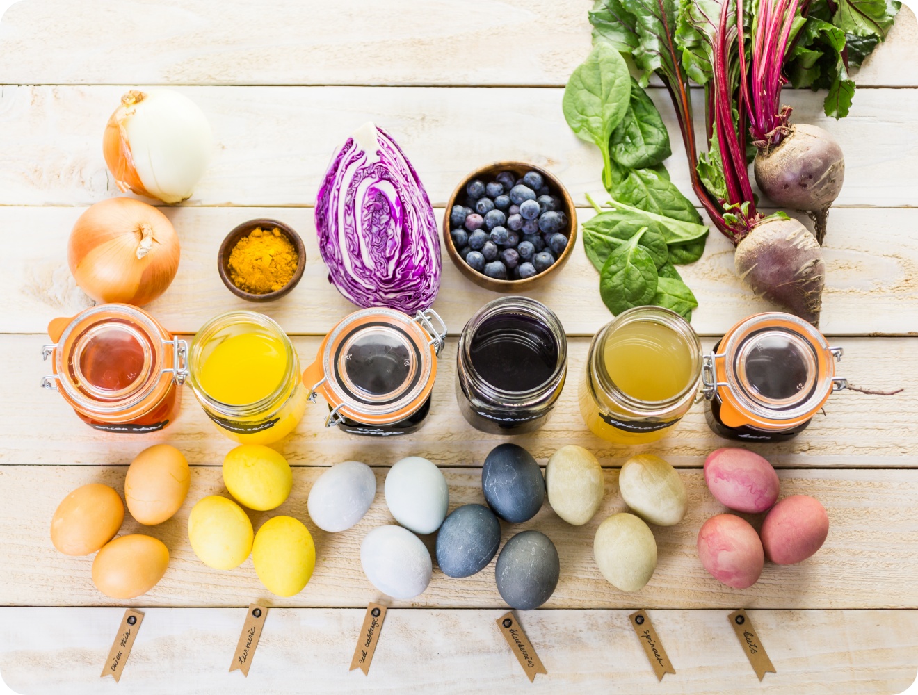 Natürliche Farbsude in Vorratsgläsern aus Zutaten wie Kurkuma, Rotkohl und Rote Bete mit gefärbten Eiern auf einem Holztisch arrangiert.