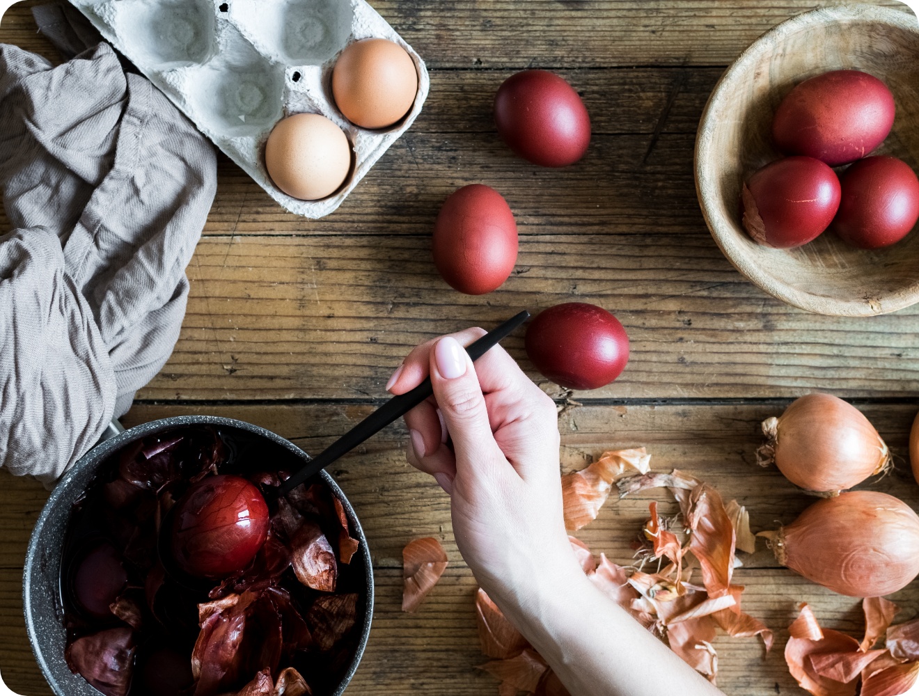 Person hebt ein tiefrot gefärbtes Osterei aus einem Topf mit Zwiebelschalen, umgeben von Eiern und Schalen auf Holztisch.