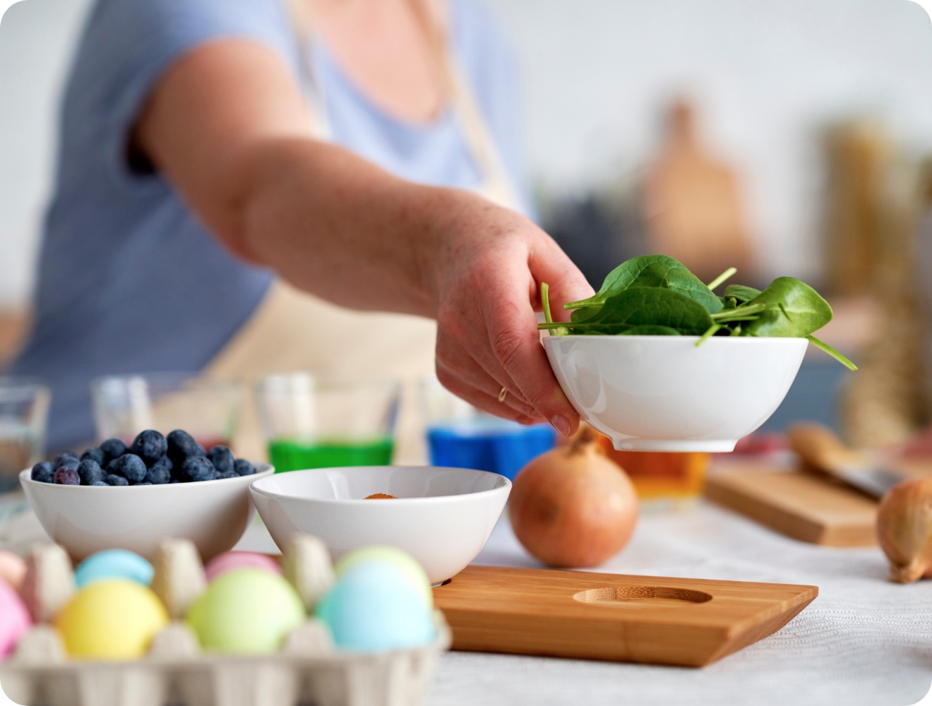 Person reicht eine Schüssel mit frischem Spinat. Auf dem Tisch stehen Blaubeeren, Zwiebeln und bunt gefärbte Ostereier.
