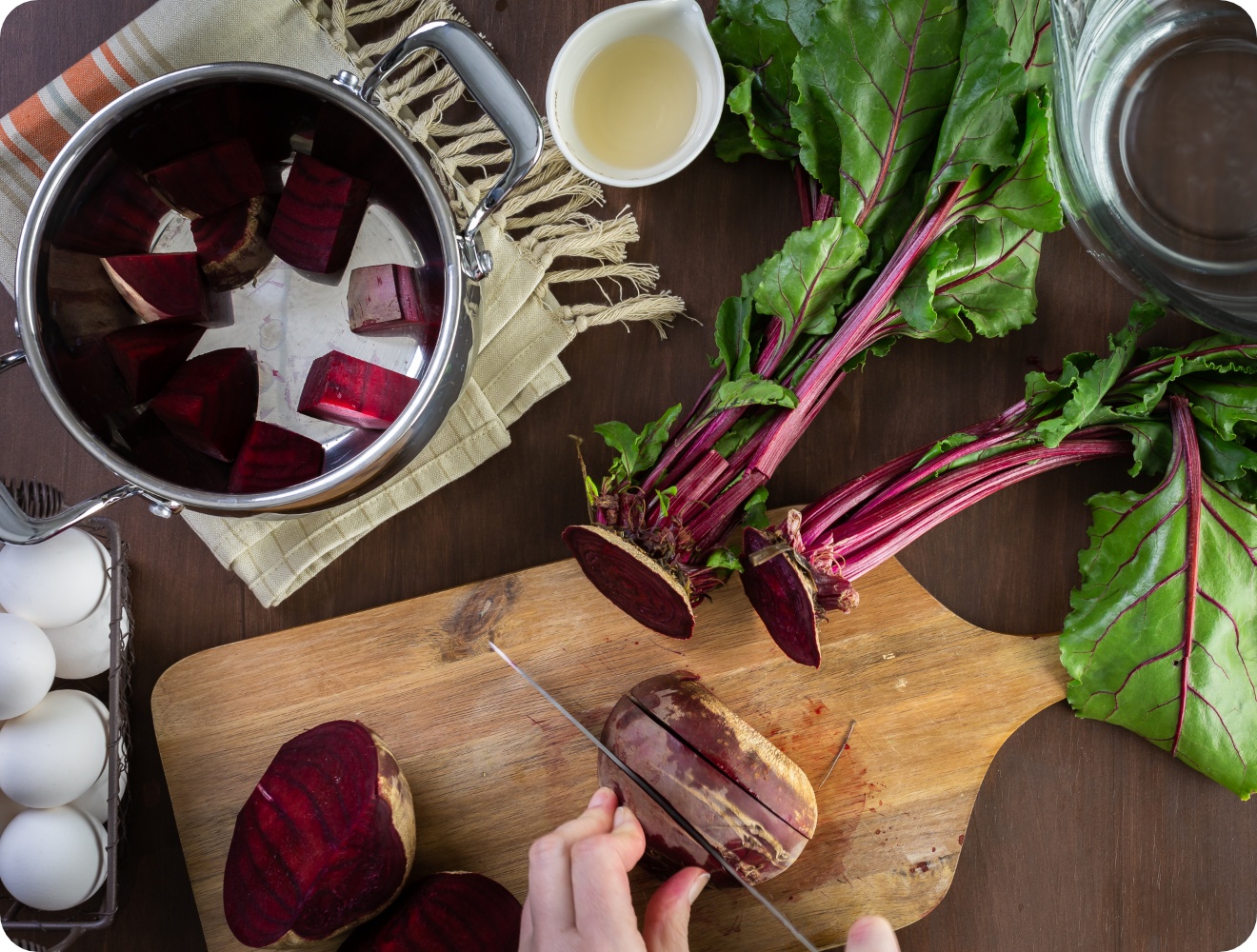 Person schneidet frische Rote Bete auf einem Holzbrett und gibt sie in einen Topf mit Essig. Auf dem Tisch stehen weiße Eier.