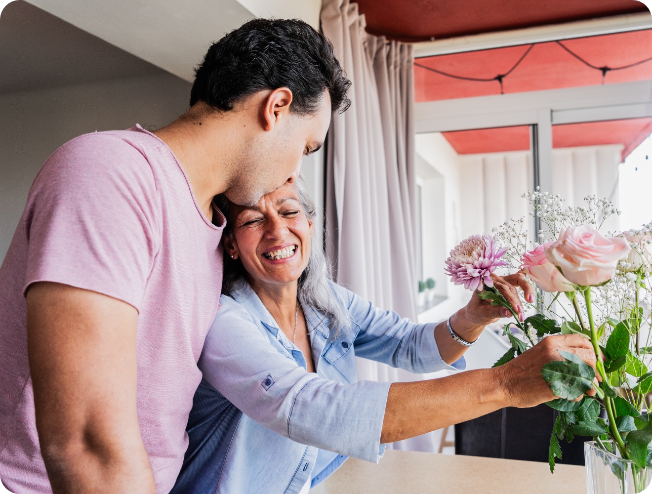 Erwachsener Sohn umarmt seine Mutter beim Blumenschenken, beide lächeln herzlich in heller Wohnung.