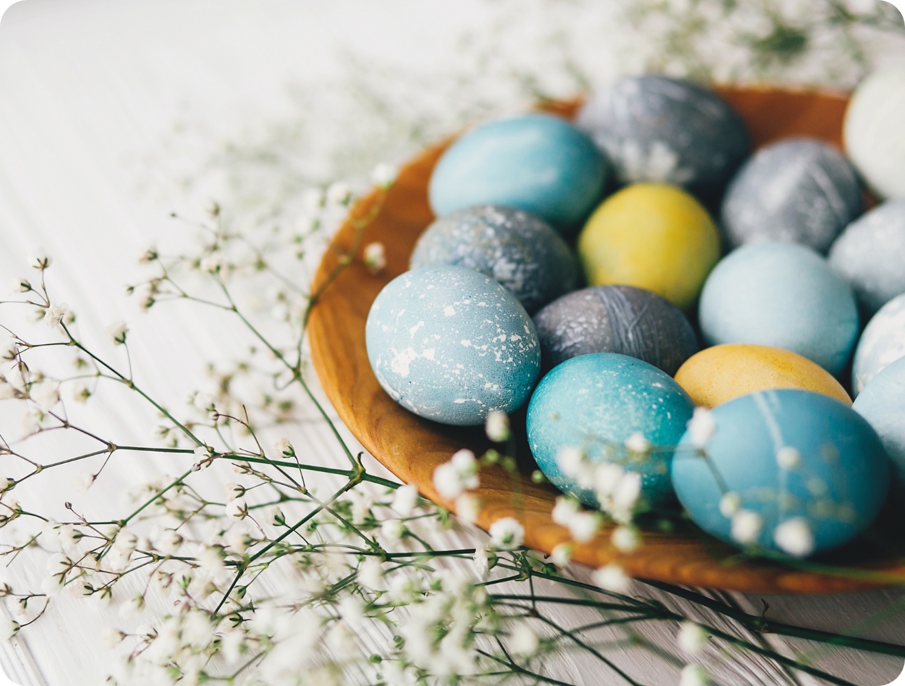 Bunter Ostereier-Mix in Blau, Gelb und Grau auf einem Holzteller, dekoriert mit zarten weißen Blüten.