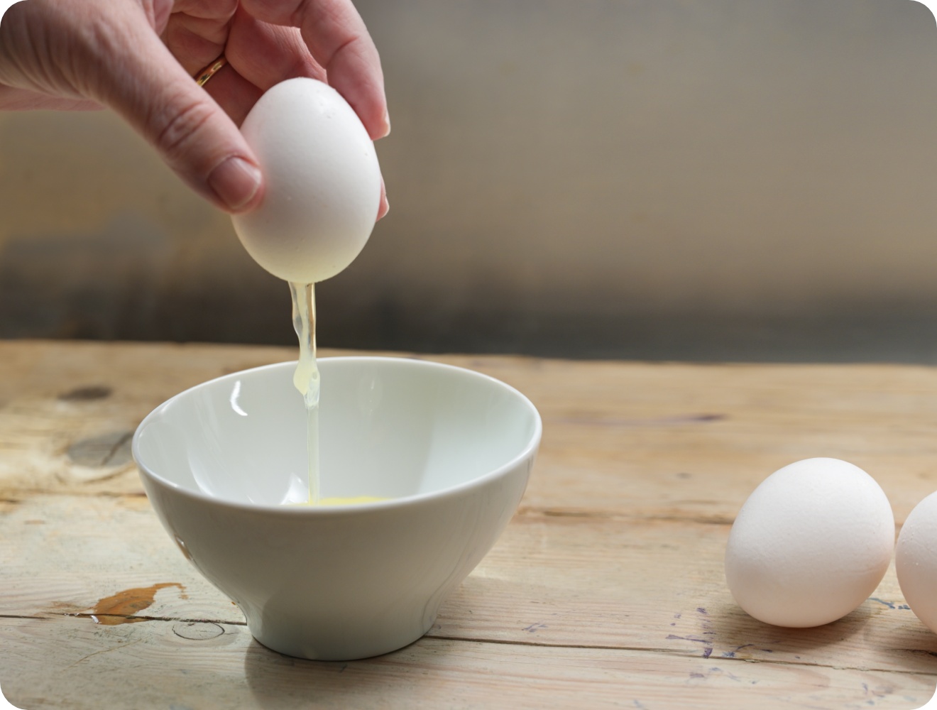Eine Person bricht ein Ei über einer weißen Schüssel auf, während das Eiweiß hinabfließt. Zwei ganze Eier liegen daneben.