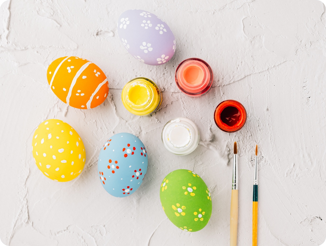 Bunt bemalte Ostereier mit Blumenmustern, Pinsel und Farbtöpfe auf strukturiertem weißen Hintergrund.