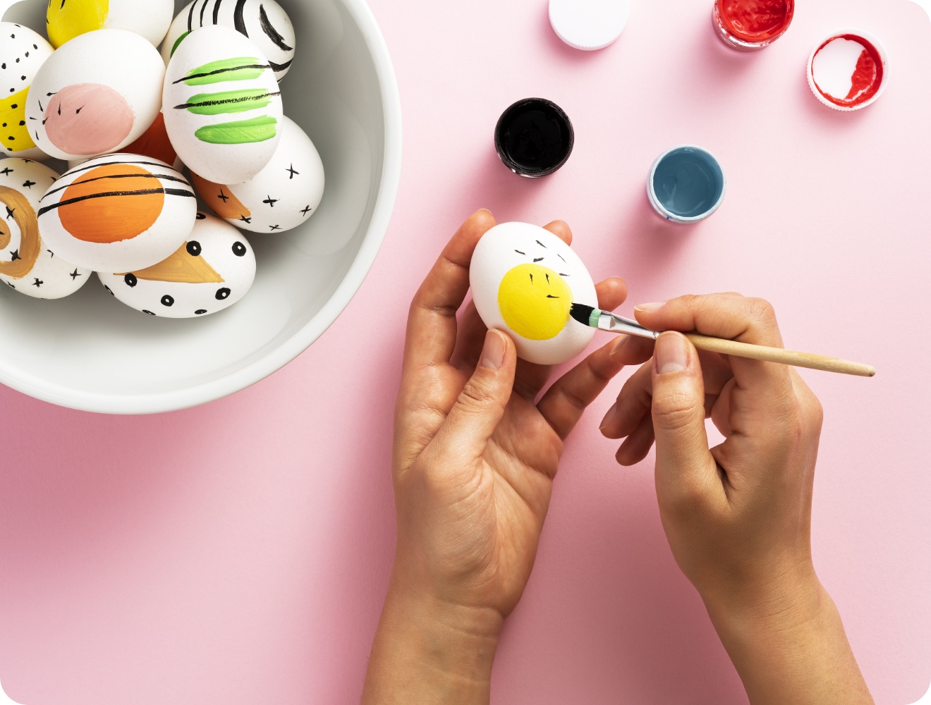 Person bemalt ein Osterei mit gelbem Kreis und schwarzen Strichen, daneben eine Schüssel mit modern verzierten Eiern.