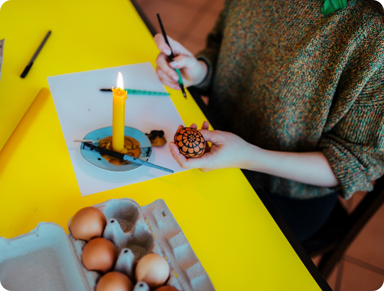 Person gestaltet ein Osterei mit Wachsbatik-Technik, gelbe angezündete Kerze steht auf dem Tisch.