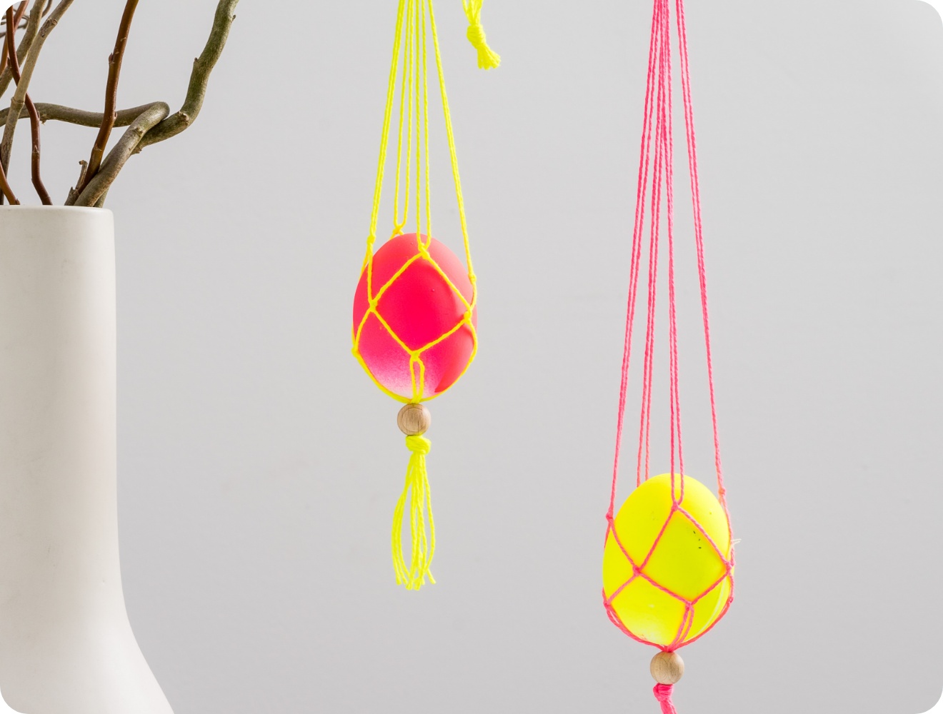 Bunte Ostereier in Makramee-Netzen aus gelben und pinken Fäden, hängend an Frühlingszweigen