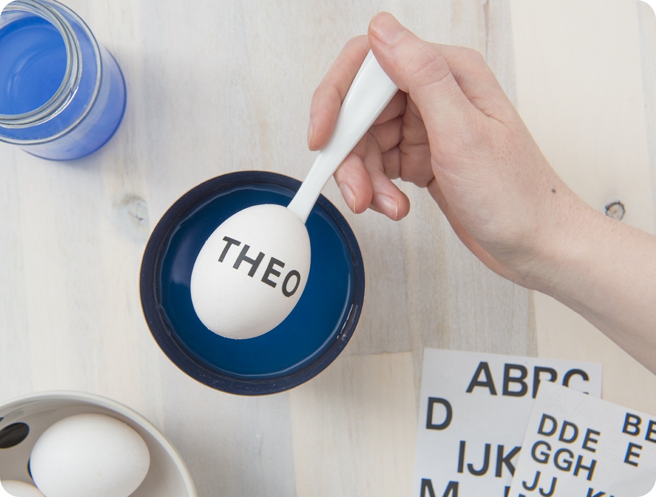Weiße Hand taucht ein Osterei mit THEO-Schriftzug in blaue Farbe, daneben Klebebuchstaben und ein Glas mit blauer Flüssigkeit.