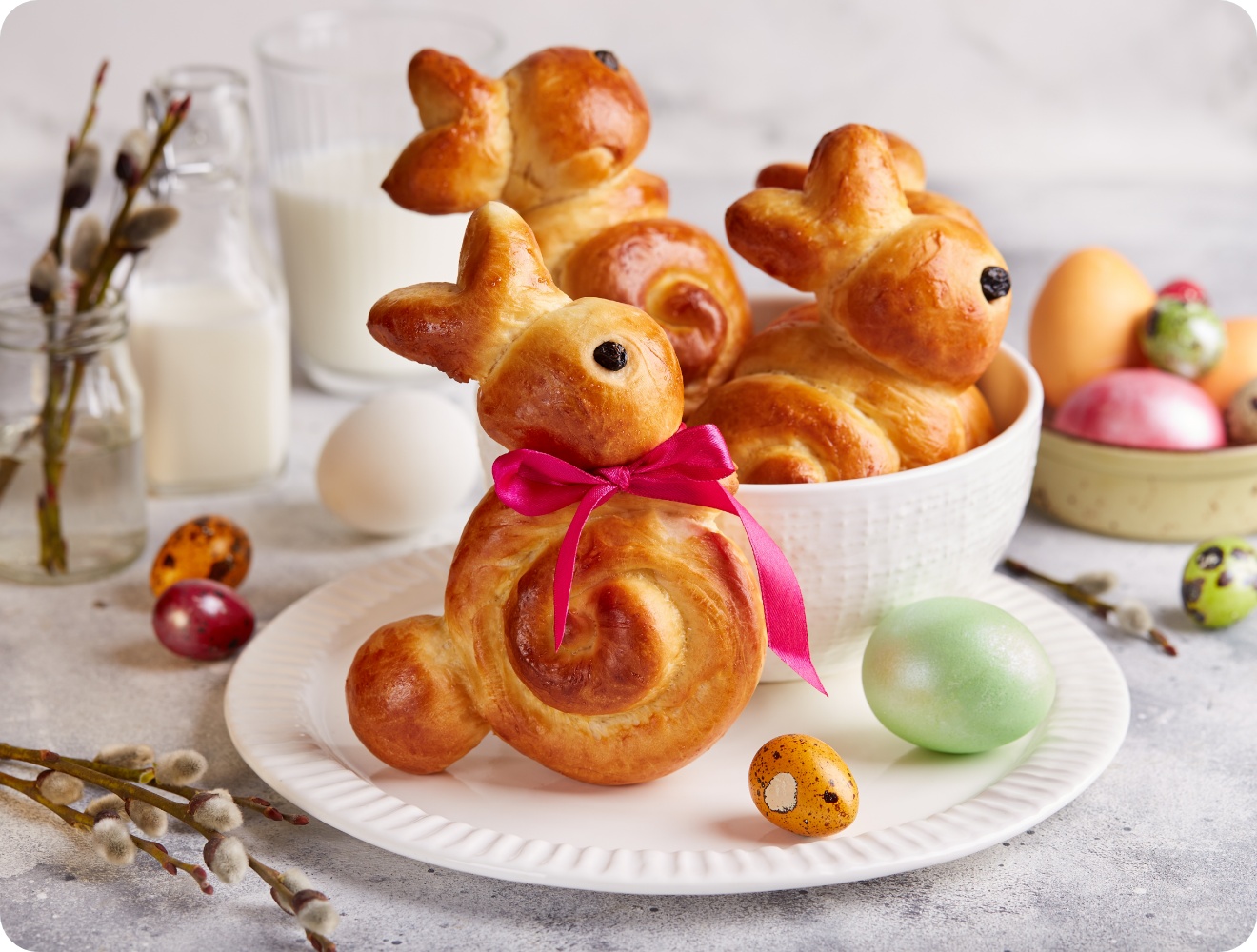 Hefegebäck in Hasenform mit Schleife, serviert zu Ostern mit bunten Eiern und Milch im Hintergrund.