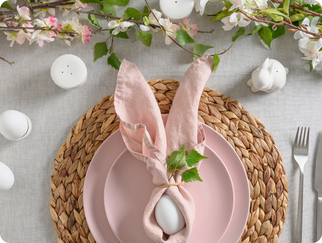 Oster-Tischdekoration mit rosafarbenem Geschirr, Ei in Serviette als Hasenohren gefaltet und blühenden Zweigen.