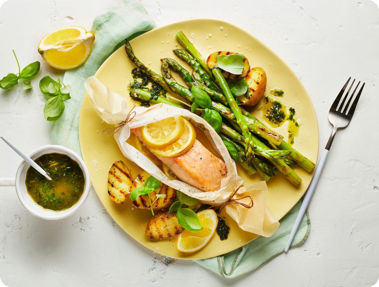 Lachs im Pergament mit Zitronenscheiben, gegrilltem Spargel und Kartoffeln auf einem gelben Teller, serviert mit Kräutersauce.