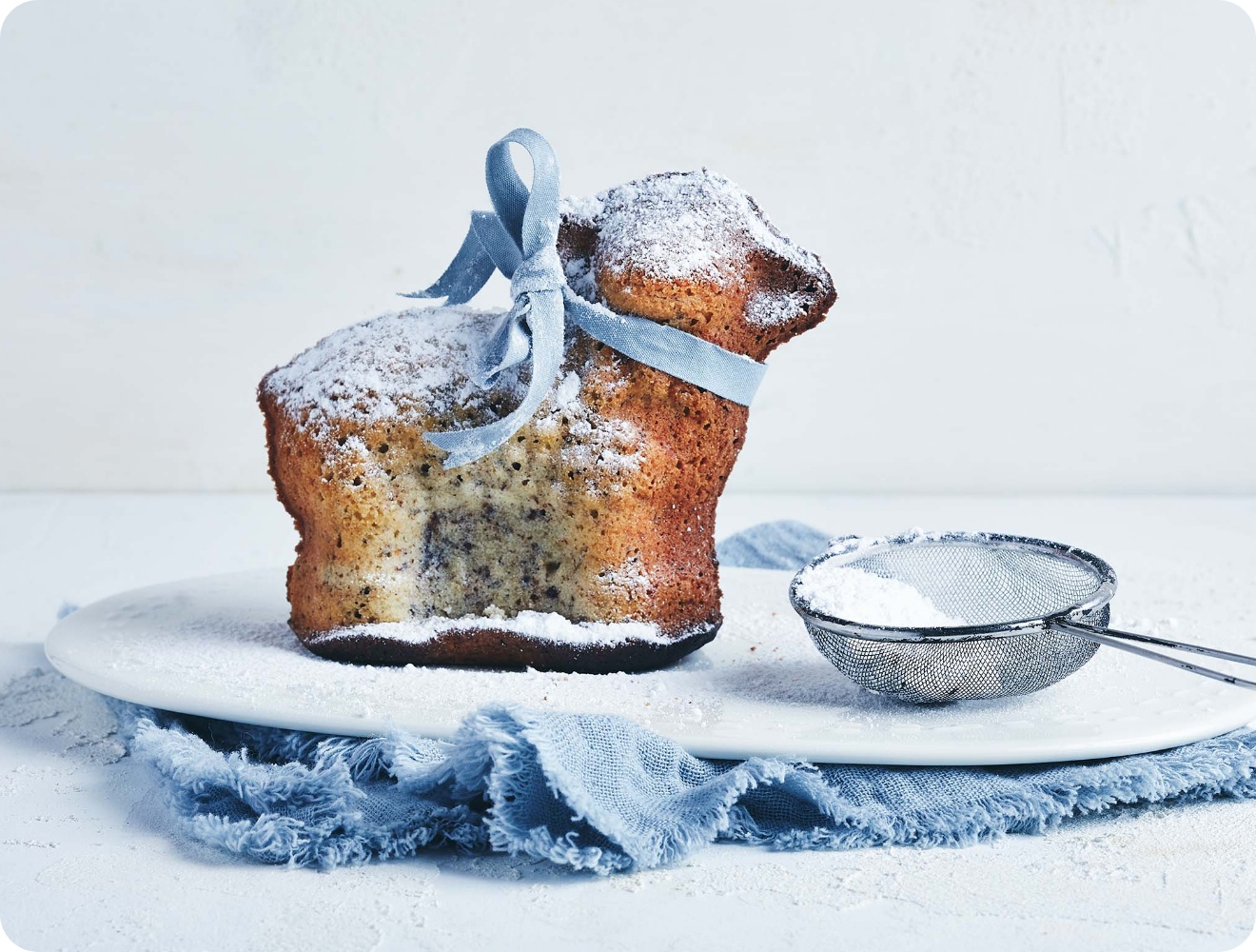 Gebäck in Lammform mit Puderzucker bestäubt, verziert mit blauer Schleife, auf weißem Teller mit Sieb und blauer Stoffserviette.