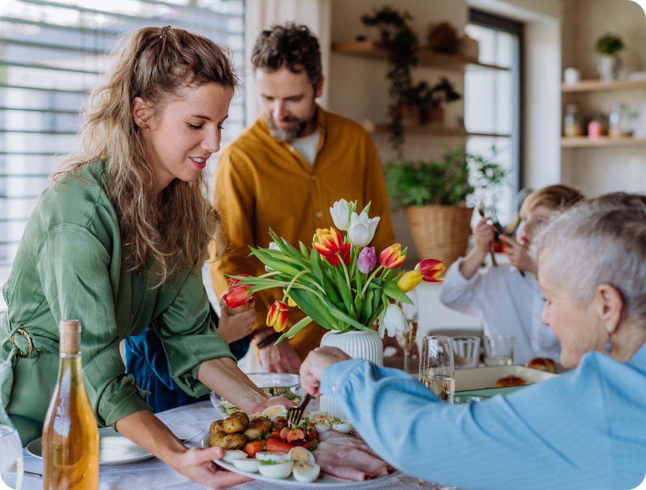 Familie genießt gemeinsames Essen an festlich gedecktem Tisch mit Tulpenstrauß, während eine Frau ein Gericht serviert.