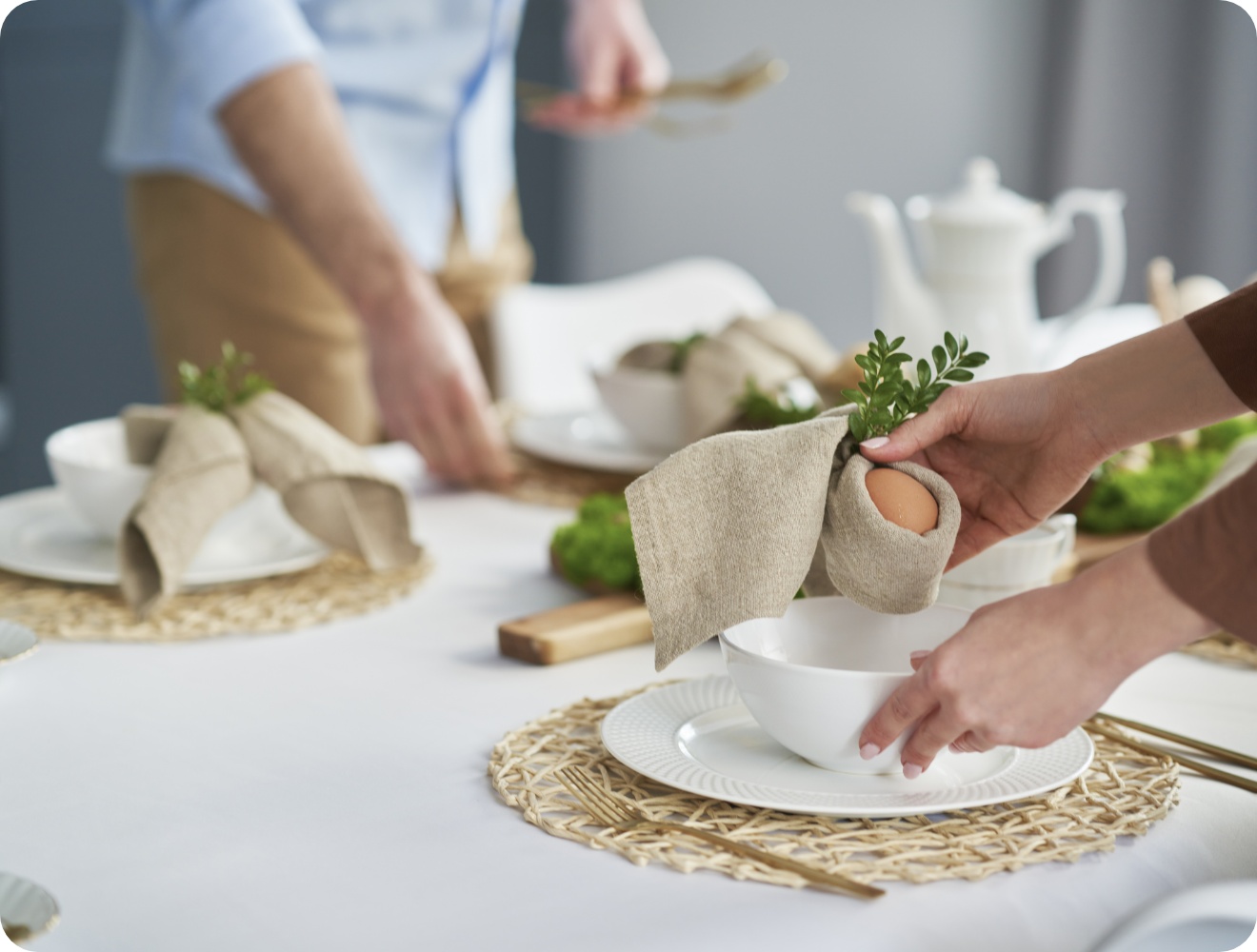 Festlich gedeckter Tisch mit handgefalteter Serviette, die ein Ei und Grünzweig umhüllt, als Osterdekoration auf einem weißen Teller.