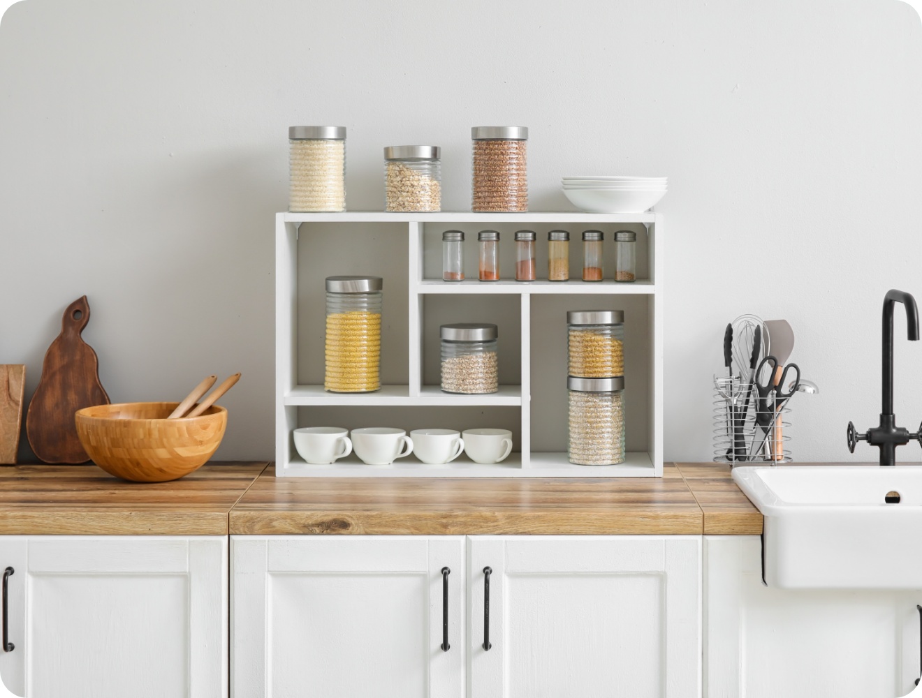Minimalistische Küche mit weißem Regal, gefüllt mit Vorratsgläsern, Gewürzen, Tassen und Schüsseln auf Holzarbeitsplatte.