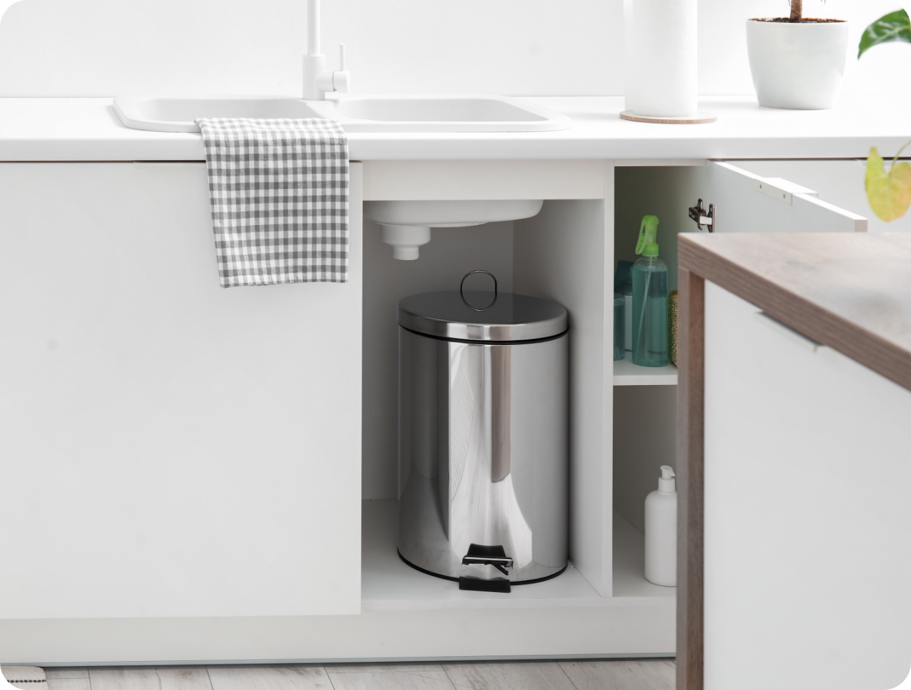 Geöffneter Küchenschrank unter der Spüle mit Edelstahl-Mülleimer und Putzmitteln in einer weißen, modernen Küche.