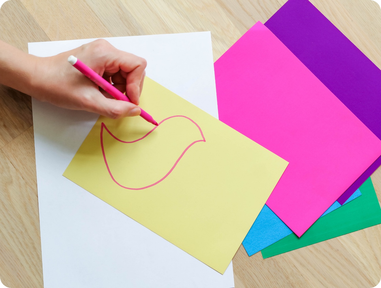 Hand zeichnet mit pinkem Stift eine Vogelkontur auf gelbes Bastelpapier. Bunte Papierbögen liegen auf einem Holztisch.