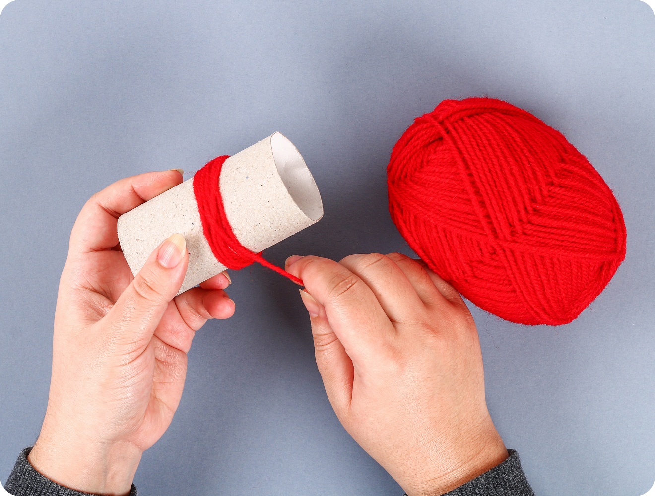 Hände wickeln rote Wolle um eine Papprolle, daneben ein Wollknäuel auf grauem Hintergrund. DIY-Bastelprojekt.