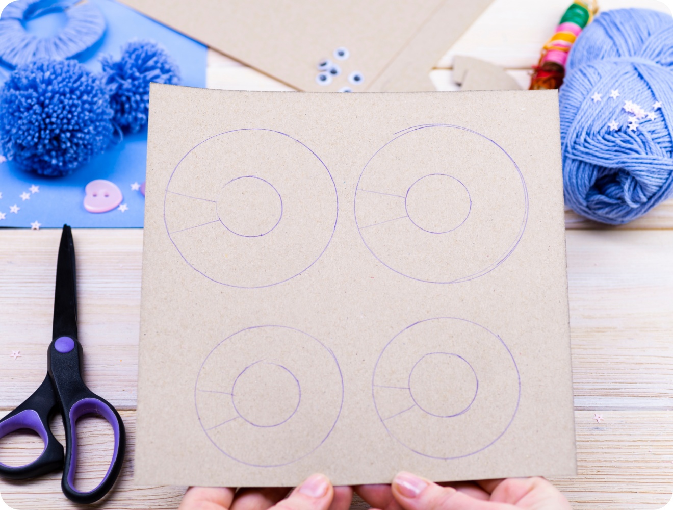 Pappscheibe mit Kreisen, Schere, blaue Wolle und fertige Pompons auf Holztisch.