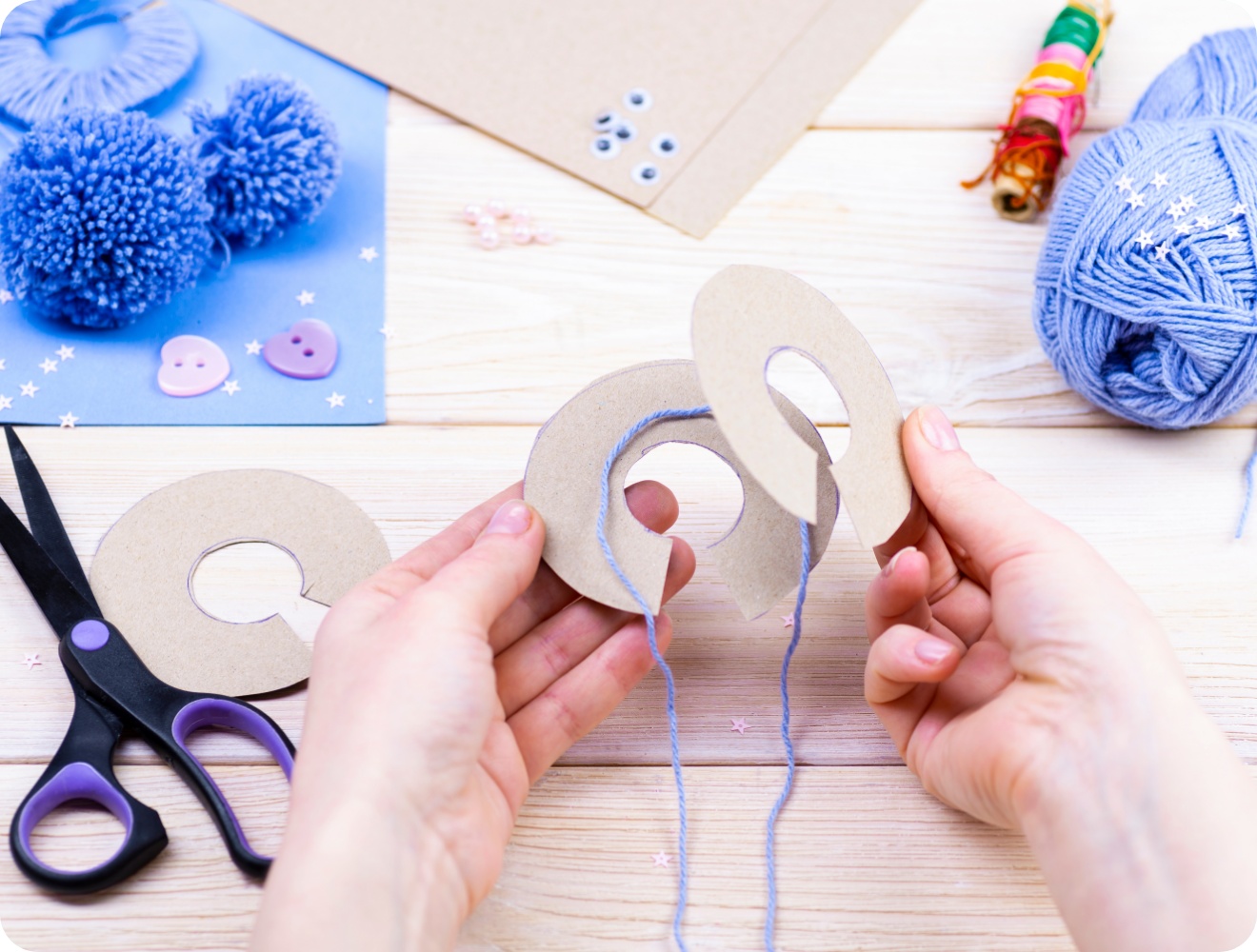 Hände wickeln blaue Wolle um Pappschablonen zur Herstellung von Pompons. Bastelmaterialien auf hellem Holztisch.