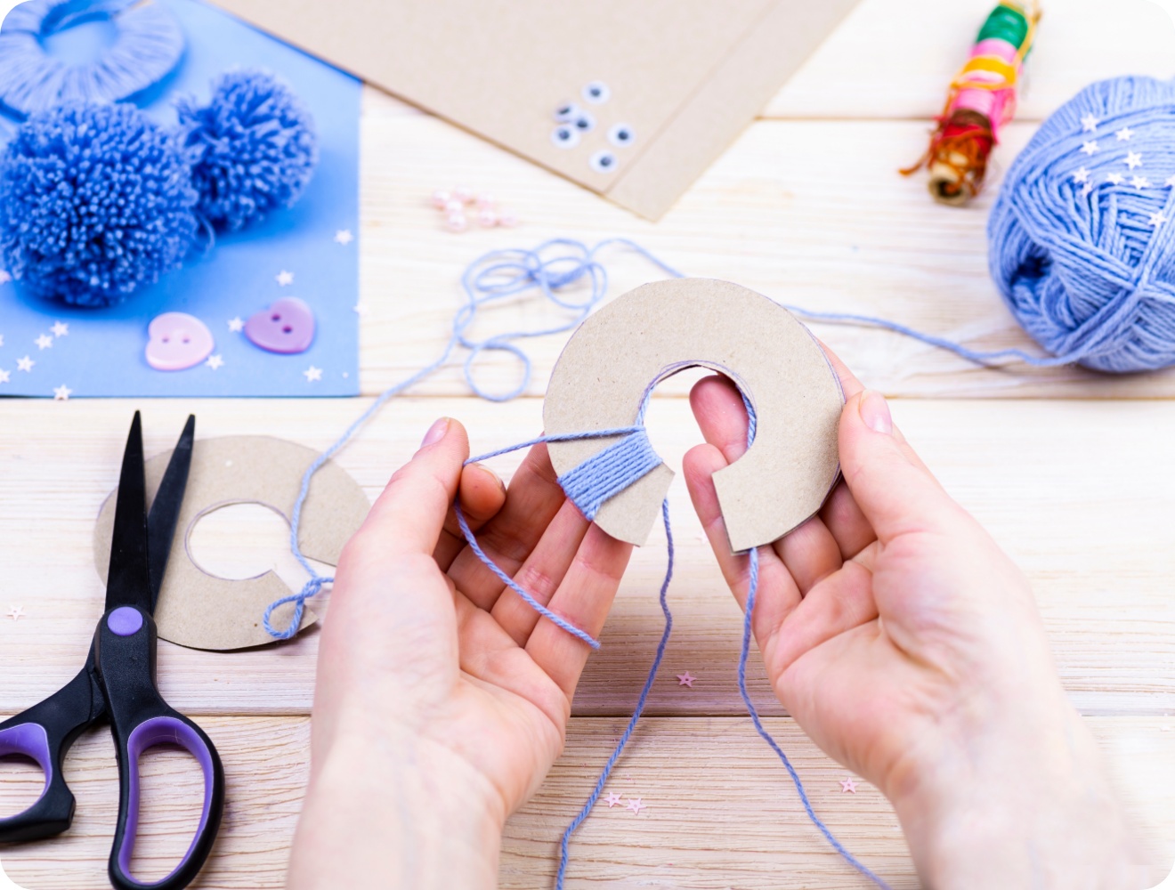 Hände wickeln blaue Wolle um eine Pappschablone für Pompons. Bastelmaterialien wie Schere und Knöpfe auf Holztisch.