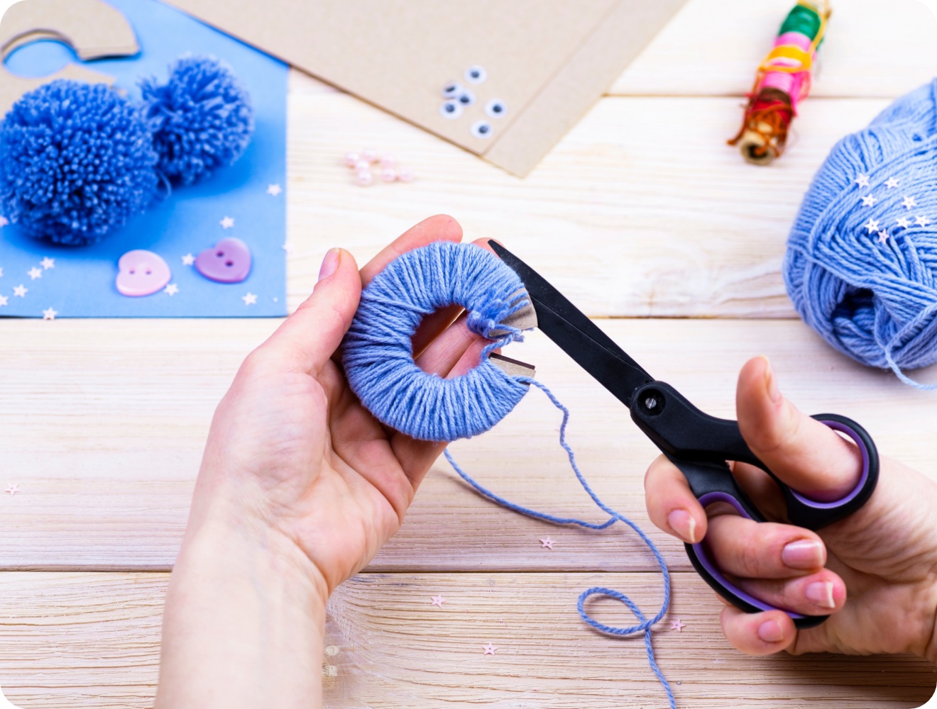 Hände schneiden blaue Wolle auf einer Pompon-Schablone. Bastelmaterialien wie Garn und Schere auf hellem Holztisch.