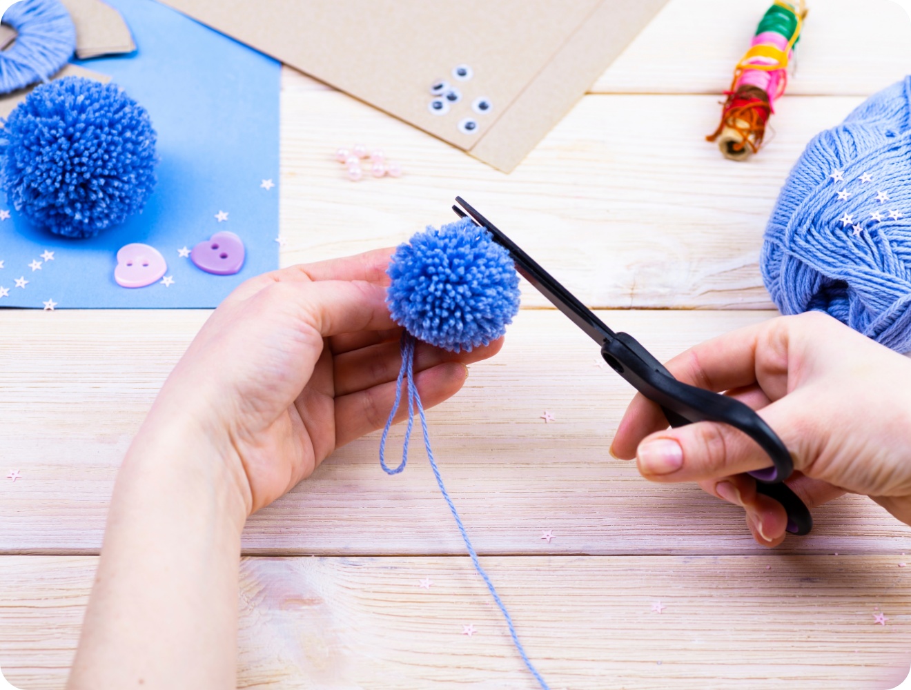 Hände schneiden mit einer Schere einen blauen Woll-Pompon zurecht. Bastelmaterialien auf hellem Holztisch.