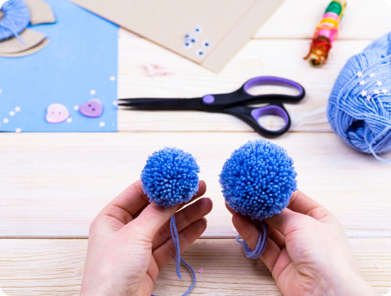 Hände halten zwei unterschiedlich große blaue Woll-Pompons. Bastelmaterialien wie Schere und Garn liegen auf einem Holztisch.