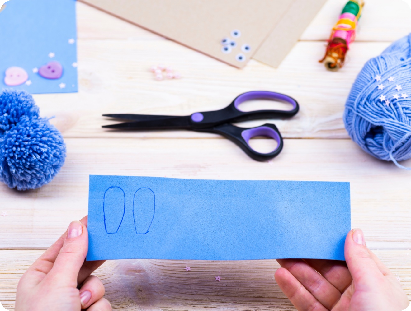 Hände halten blaues Bastelpapier mit aufgezeichneten Hasenohren. Bastelmaterialien wie Schere, Wolle und Pompon liegen auf Holztisch..