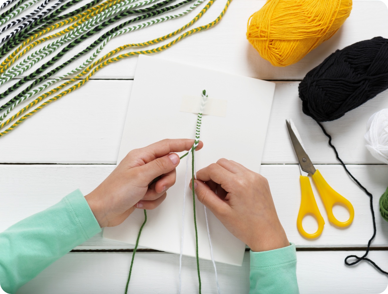 Hände flechten ein Freundschaftsarmband aus grüner, weißer und gelber Wolle. Bastelmaterialien auf weißem Holztisch.