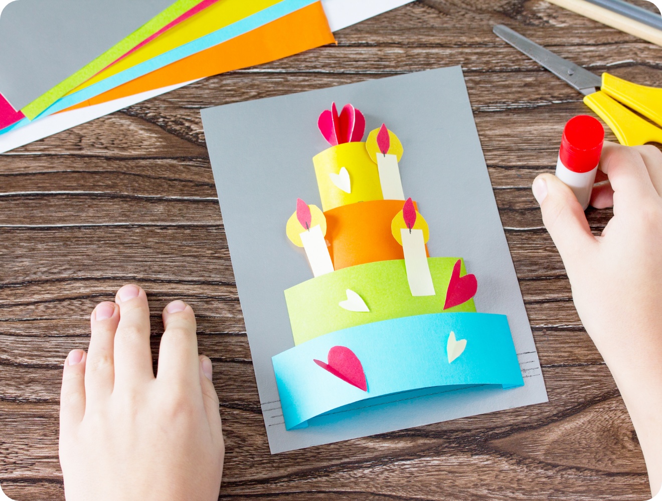Kinderhände basteln eine bunte 3D-Geburtstagskarte mit einer Papier-Torte und Kerzen. Bastelmaterialien auf Holztisch.