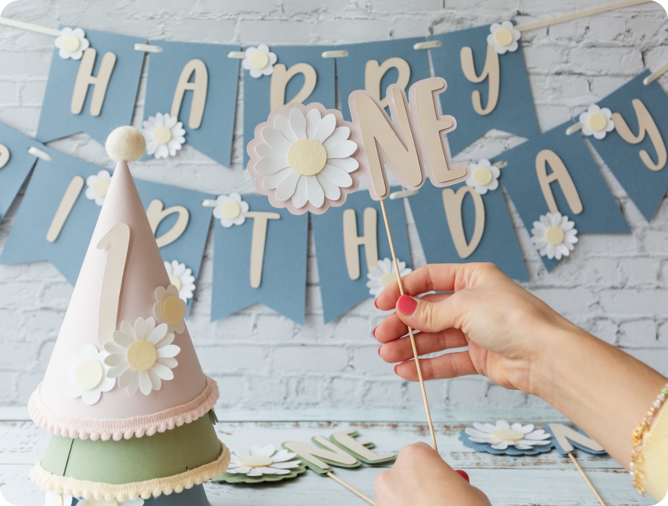 Hand hält eine DIY-Geburtstagsdekoration. Im Hintergrund blaue Happy-Birthday-Girlande und Partyhut.